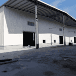 The exterior of a white metal structure barn building has large open doors, a slanted roof, and concrete ground in front.