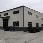 A beige Metal Structure Barn Building with blue trim, multiple windows, and a large entrance, surrounded by some materials and equipment outside.