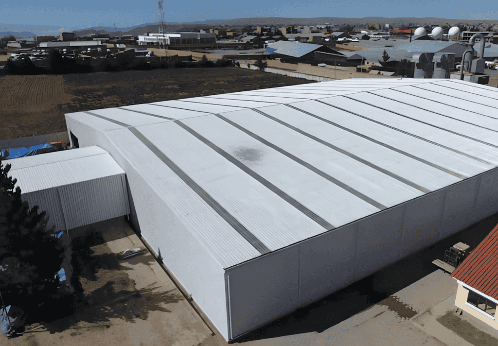 Aerial view of a white Metal Structure Barn Building with a patterned roof, situated in an open area with other buildings and greenery in the background.