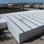 Aerial view of a white Metal Structure Barn Building with a patterned roof, situated in an open area with other buildings and greenery in the background.