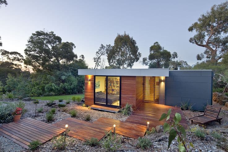 Modern modular home with stylish landscaping, wooden deck, and ambient outdoor lighting in a natural setting