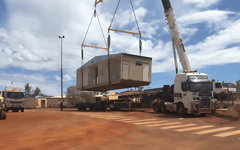 Crane hoisting a modular housing unit onto a flatbed truck, with workers assisting on-site under a bright sky at a modular construction location.