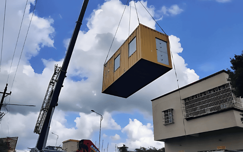 Large crane lifting a yellow container-style modular home unit into the air, with residential buildings and blue sky with clouds in the background.