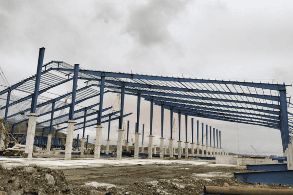 A large steel framework for an industrial building, partially completed with visible columns and beams on a cloudy day.