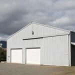 A light - gray pole barn tractor shed featuring two closed white doors, set against a cloudy sky near another building.