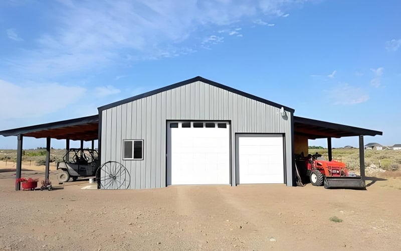 A gray pole barn tractor shed with a black roof overhang, two white garage doors, and farm equipment like a tractor parked outside.