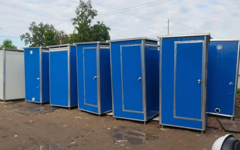 A row of blue portable toilets outdoors on a dirt - ground with trees and power lines in the background.