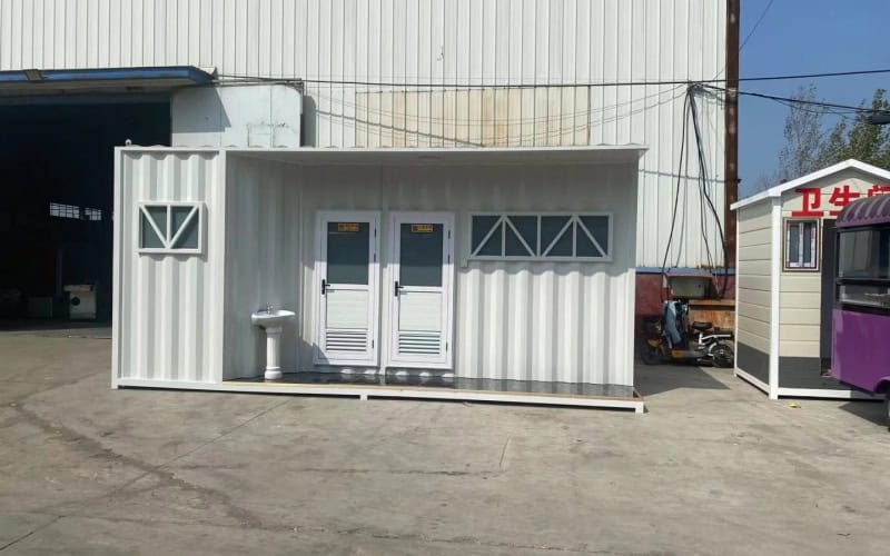 White prefab ablution block with two doors, a sink outside, and windows, situated in front of a industrial building.