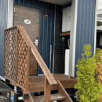 Compact modular home with blue corrugated walls, wooden stairs, and small porch surrounded by greenery for a cozy and functional outdoor feel.