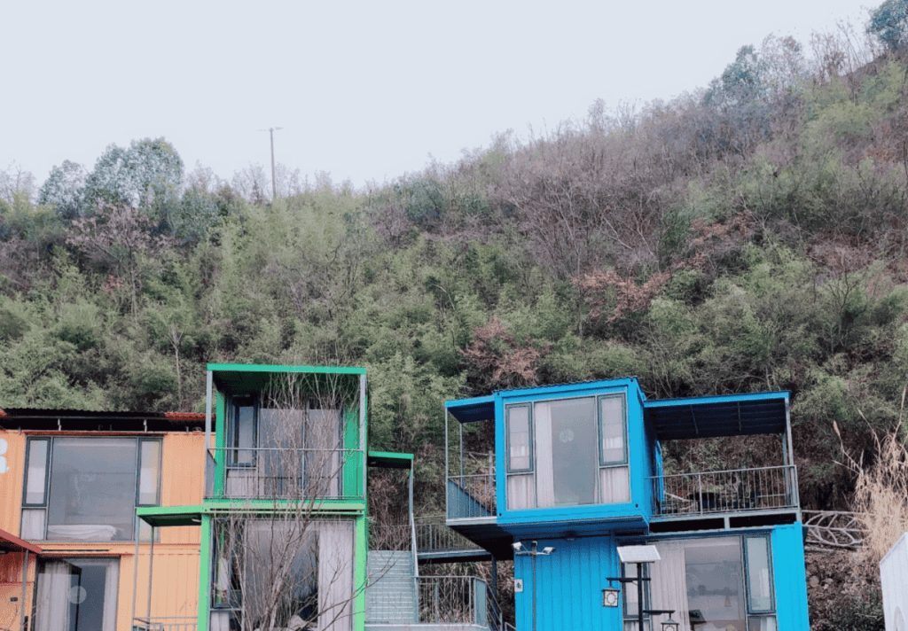 Colorful stacked modular container homes with green and blue accents against a lush hillside backdrop, creating a vibrant and eco-friendly atmosphere.