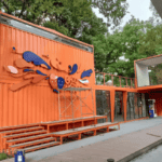 Orange modular container building with creative design, featuring a playful wall installation, large windows, and an inviting outdoor seating area.