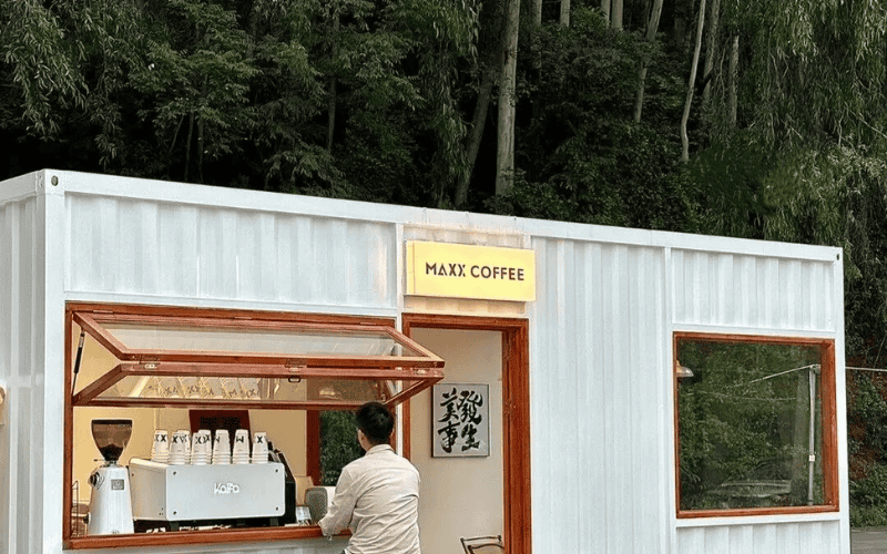 A small coffee shop in a shipping container, with a barista serving customers from a window, surrounded by greenery.