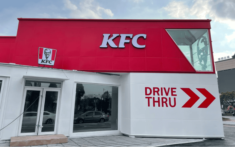 A container KFC restaurant with a bright red exterior and a large 'Drive Thru' sign on the wall, in front of a modern building.