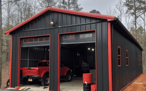 A metal garage with black and red siding, two open doors, one with a red truck parked inside, surrounded by trees.