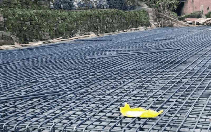 A large raft foundation reinforced with a steel mesh grid, set in a construction site surrounded by natural stone walls.