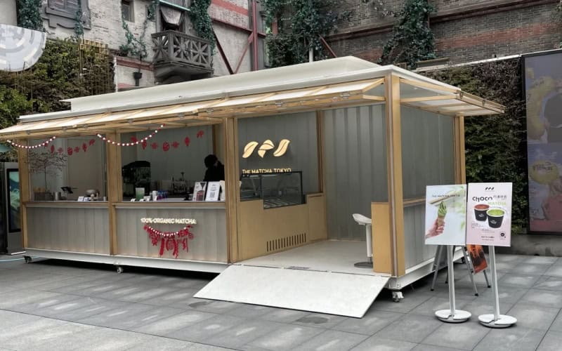 A retail container shop with a light grey and wooden exterior, featuring a canopy, festive lights. Promotional boards are placed outside, set against an urban backdrop with greenery - covered buildings.