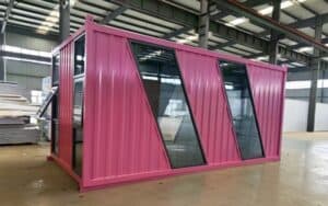A pink retail container shop with geometric - shaped glass windows, positioned inside an industrial workshop, highlighting a modern and vibrant design.