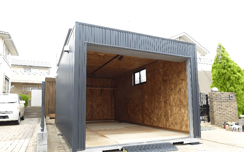 A grey shipping container garage with an open roller shutter door, revealing a plywood - lined interior, situated on a paved driveway between residential buildings, with a car and greenery in the background.