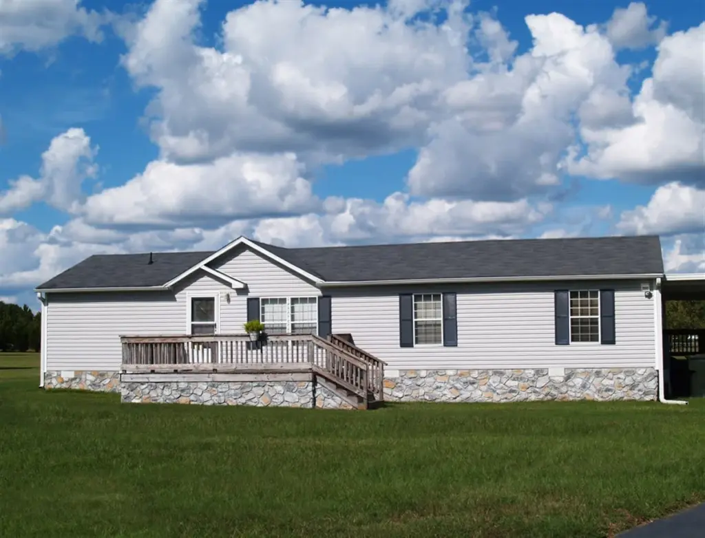 Simple modular home with dark asphalt shingle roof, vinyl siding, dark shutters, front porch, and stone skirting on a well-maintained lawn under a blue sky