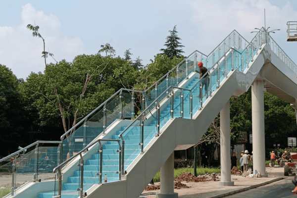 A pedestrian skybridge staircase with glass railings and a vibrant blue design, surrounded by trees.