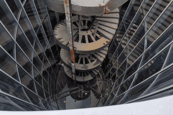 A tall steel spiral staircase surrounded by reflective glass walls, viewed from above.