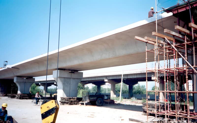 Under - construction girder bridge with concrete piers supporting a large beam, surrounded by construction materials and scaffolding.