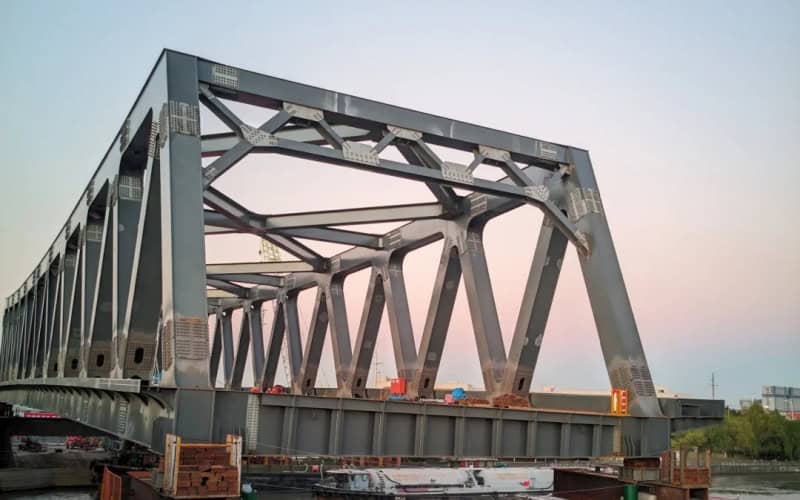 Close-up of a large steel structure bridge under construction by a waterway, with its intricate truss design visible.