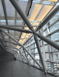 Interior of a modern glass and steel corridor with angled white beams and large windows overlooking city skyscrapers.