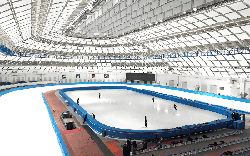 Large steel structured skating rink with a high, arched roof and skaters on the ice.