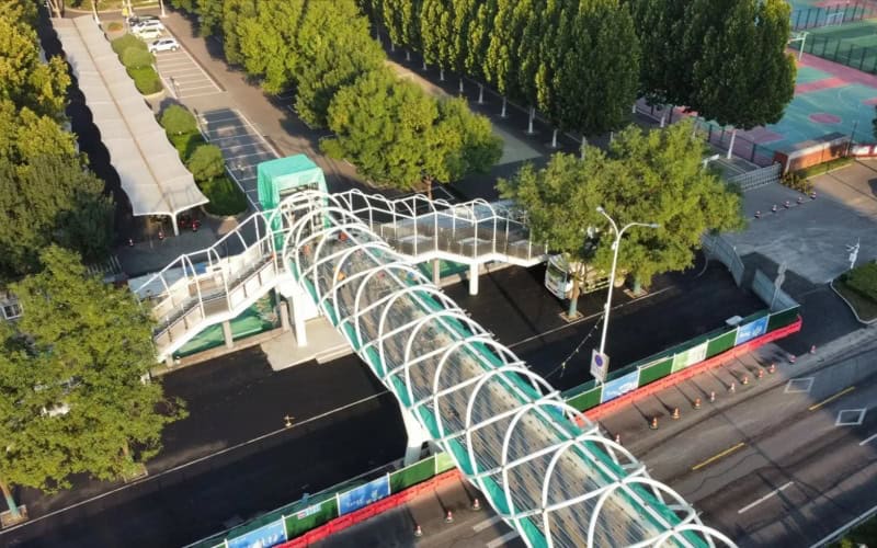 Aerial view of a steel - structure pedestrian flyover with white arches and green coverings, spanning over a road with trees and a parking area nearby.