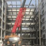 A red crane hoisting materials for a multi-story Steel Structure Office Building under construction between two existing buildings.