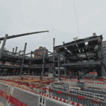 Construction scene of a Steel Structure School Building, with a crane hoisting components. Safety fences and workers are visible around the building site.