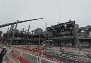 Construction scene of a Steel Structure School Building, with a crane hoisting components. Safety fences and workers are visible around the building site.