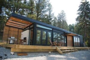 Stylish modern modular home with black metal framing, floor-to-ceiling windows, wood accents, and a spacious deck in a forest setting