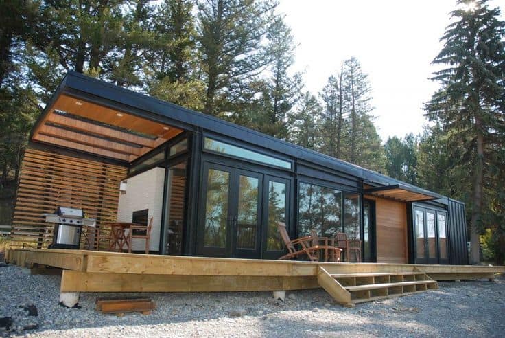 Stylish modern modular home with black metal framing, floor-to-ceiling windows, wood accents, and a spacious deck in a forest setting