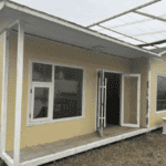 A front view of a yellow prefab house with a covered porch and large windows, designed for comfort and practicality in an open area.