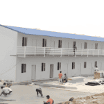 A two-story prefab housing complex under construction, with a blue roof and workers preparing the foundation on-site.