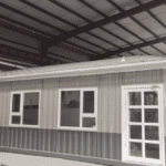 A single-story Temporary Modular Building with metallic siding, multiple windows, and glass-paneled doors, situated inside an industrial shed.