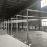 Frame structure of a Temporary Modular Building being assembled inside a spacious industrial hall, with construction materials like panels and tools scattered on the floor.