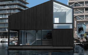 A modern modular building on water with dark panels and large glass windows, set against urban buildings.