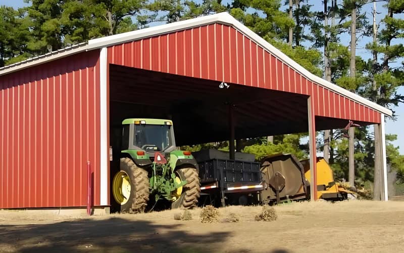 A red metal tractor shed with a white - trimmed edge, where a green tractor and farming equipment are parked inside, set against a backdrop of trees.