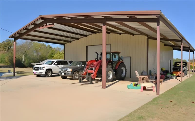 A large metal tractor shed with a brown - roofed canopy, housing a tractor, two cars, and various items on a concrete ground.