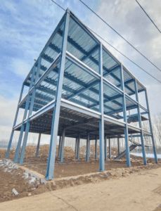 A newly constructed steel building frame with blue supports and metallic flooring, situated on a cleared plot under a blue sky.