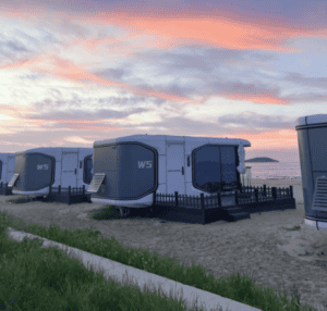 Una hilera de modernas casas cápsula ubicadas junto a la playa, con una pintoresca puesta de sol de fondo, que ofrecen una experiencia de vida única y futurista.