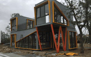 A modern two-story home constructed from shipping containers, featuring bold orange accents, large glass windows, and an industrial design.