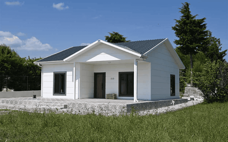 A small white prefabricated house with a modern design, featuring a sloped roof and surrounded by greenery under a blue sky.