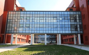 A modern steel structure school building with large glass windows and red hued walls, featuring an open area in front.