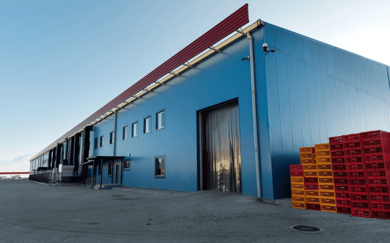 Blue hued PEB cold storage building design with large door, small windows, and red topped roof, pallets outside.