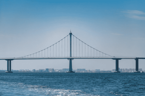 A long suspension bridge spanning across a wide body of water, with a city skyline in the background.