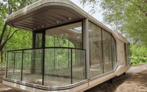 Modern prefab house with large glass windows and a balcony, surrounded by greenery, showcasing a minimalist design.